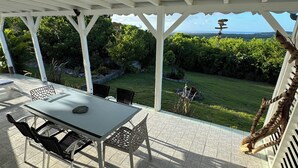 A covered patio with a table and chairs, a view of a lush garden, and a distant landscape. - Sugar Cane House (GRAND BOURG)