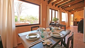 A dining area with a wooden table set for four, large windows, and a view of greenery outside. - Sa Torre: Idyllic estate, individual areas, garden, pool, WiFi, air conditioning (Santa Maria del Cami)