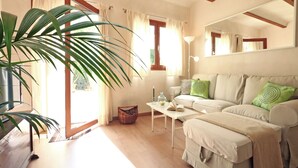 A living room with a sofa, a coffee table, a lamp, a large plant, and a window with curtains. - Sa Torre: Idyllic estate, individual areas, garden, pool, WiFi, air conditioning (Santa Maria del Cami)