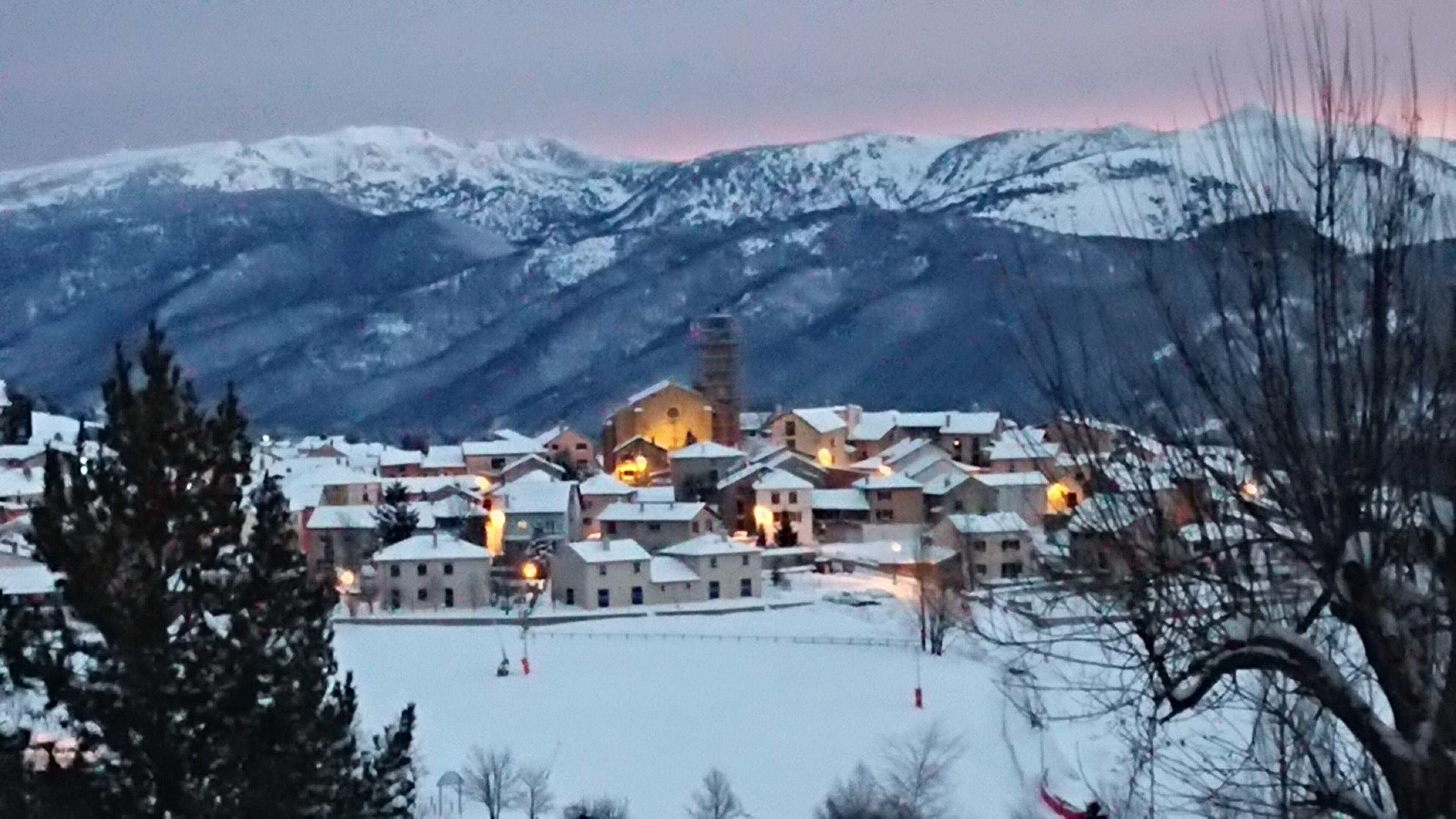 Ein schneebedecktes Dorf mit einer markanten Kirche im Zentrum, umgeben von Wohnhäusern und Bergen im Hintergrund.