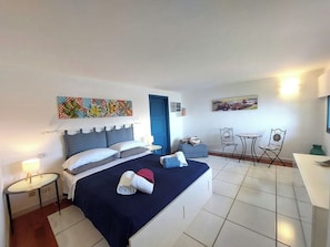 Une chambre d’hôtel comprenant un lit, un bureau, deux chaises, une petite table, une porte et des œuvres d’art accrochées aux murs.