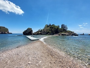 A pebble beach leading to a rocky island with clear water and a pier.