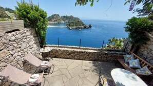 A patio with stone walls, a wooden deck, and outdoor furniture overlooking a coastal view with a rocky island and clear blue sea. - Villette Taormina, La Perla Dell'isola (Taormina)