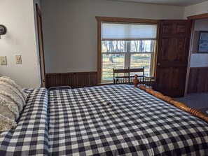 A bedroom with a checkered bedspread, a wooden headboard, a window with a view of trees, and a door to the right.