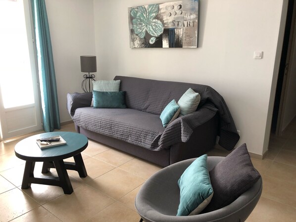 Een woonkamer met een grijze bank, kussens in teal-tint, een grijze fauteuil en een kleine ronde tafel met een afstandsbediening.
