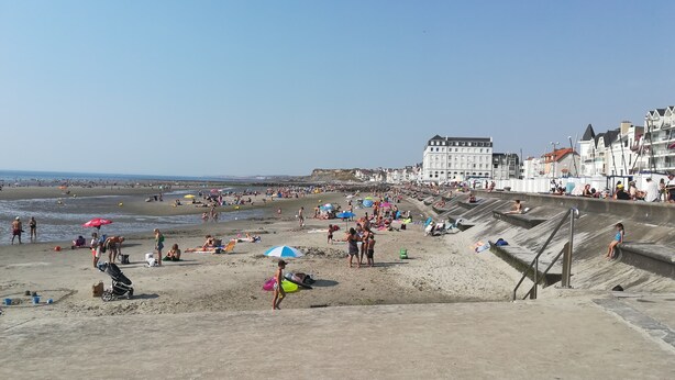 En fullsatt strand med folk som solar och badar, en stor byggnad i bakgrunden och en klar himmel.