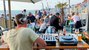 En DJ-bås med ljudutrustning, ett bord med drycker och en folkmassa på en plats vid havet.