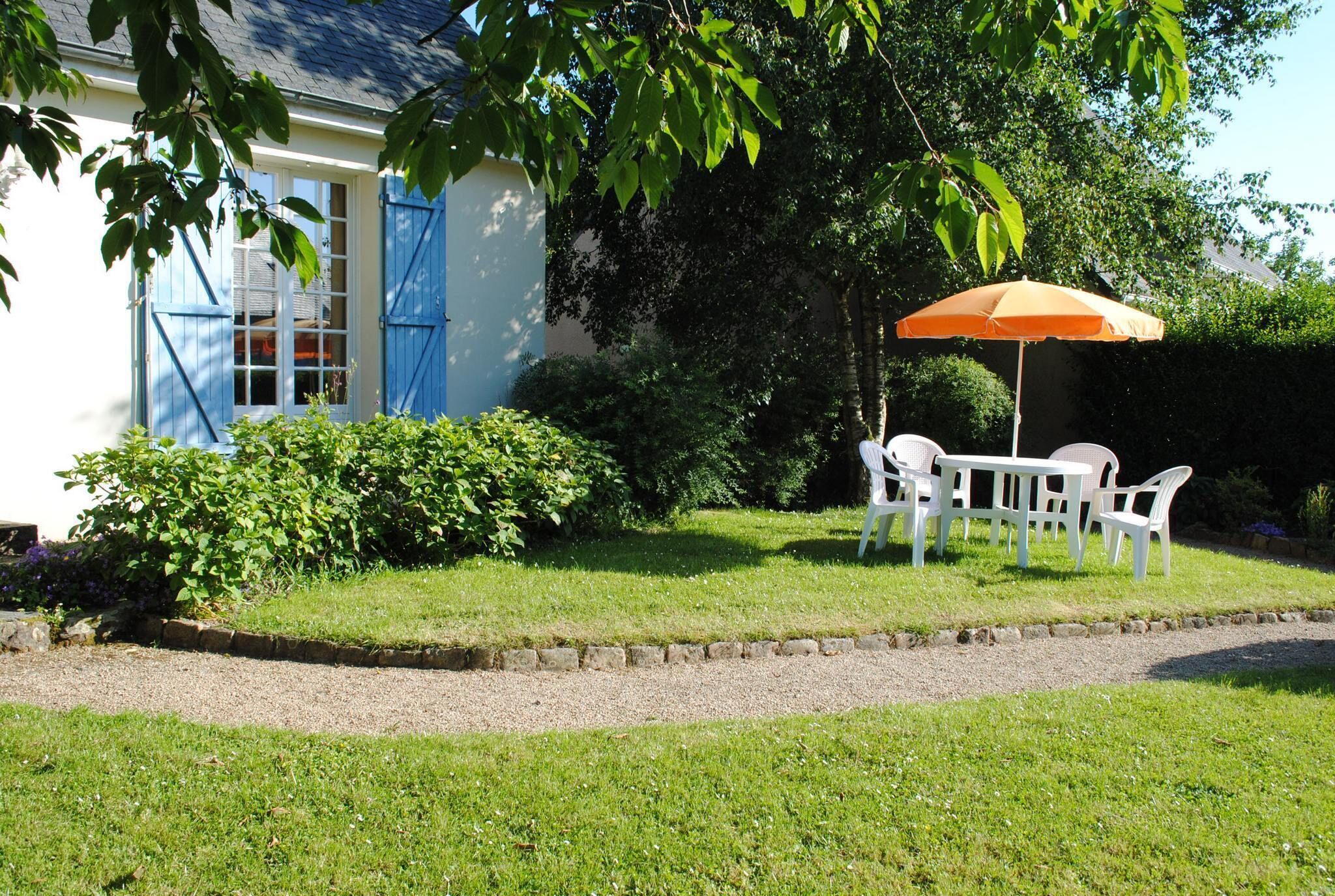 Ein Haus mit blauer Tür und Fenstern, ein Garten mit einem weißen Tisch und Stühlen unter einem orangefarbenen Sonnenschirm sowie ein Kiesweg, der zum Haus führt.