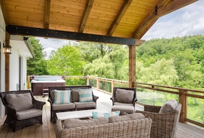 Relax on the covered deck with wicker sofas, armchairs, and a small table, overlooking a lush landscape with a hot tub in the distance.