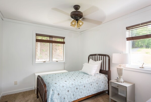 Une chambre à coucher comprenant un lit, une table de chevet, une lampe, un ventilateur de plafond et deux fenêtres équipées de stores.