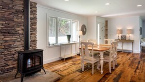 A cozy room with a stone wall, a black wood-burning stove, a dining table with chairs, and a window with a view of trees. - 36 Private Acres on the New River (Boone)
