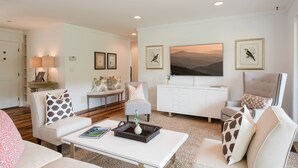 A modern living room with a white sofa, a white coffee table, and a white cabinet. There are two framed pictures on the wall, a lamp on a side table, and a painting of mountains. - 36 Private Acres on the New River (Boone)