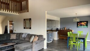 A living room with a sectional sofa, a dining area with bright green chairs, a kitchen with white cabinets, and a chandelier.