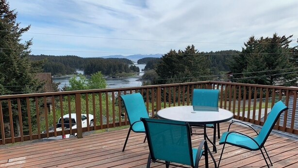 A wooden deck with a round table and four chairs, overlooking a river and forested hills.