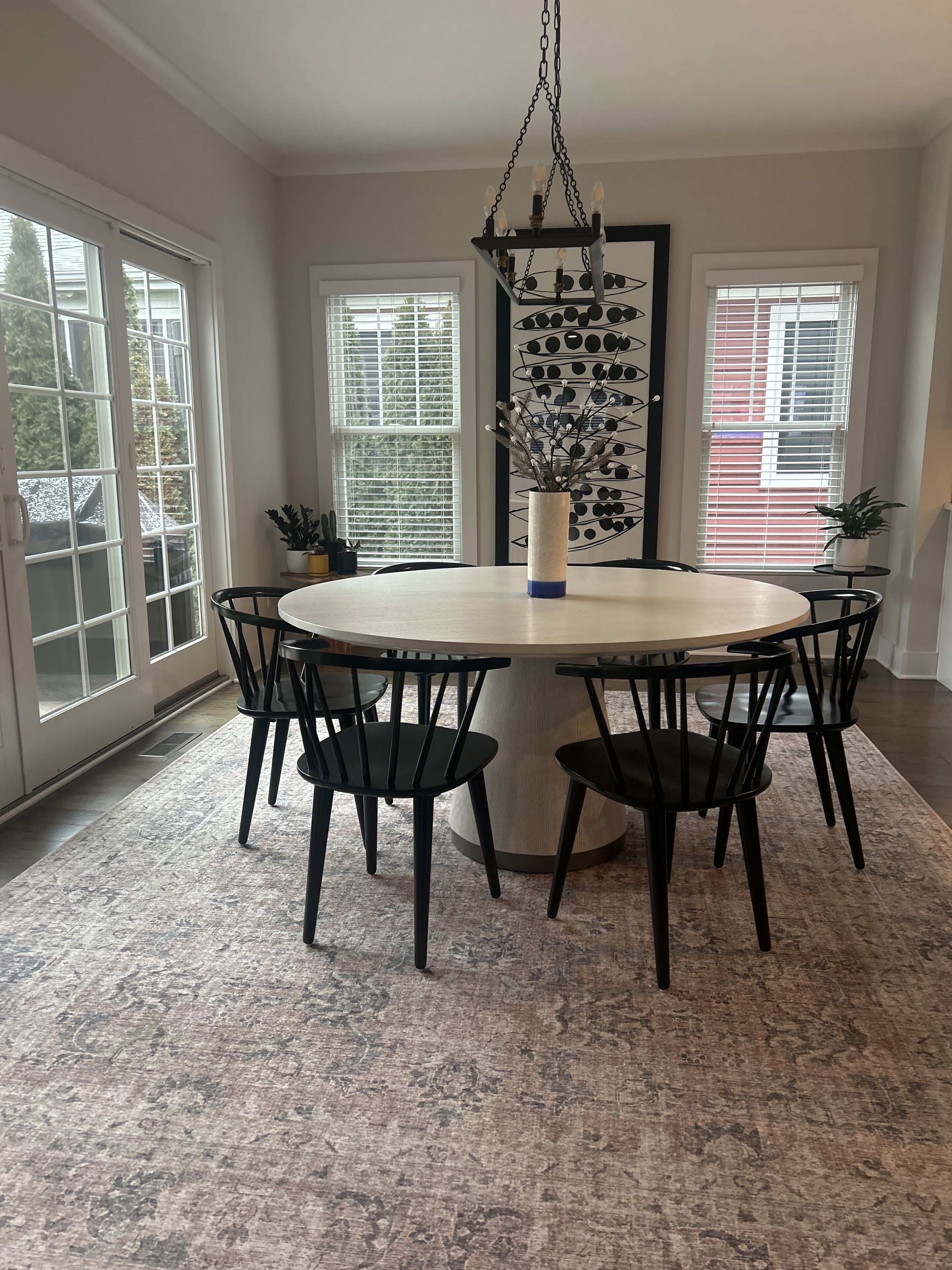 A dining room with a round table surrounded by six chairs, a hanging light fixture, and a vase with dried flowers.