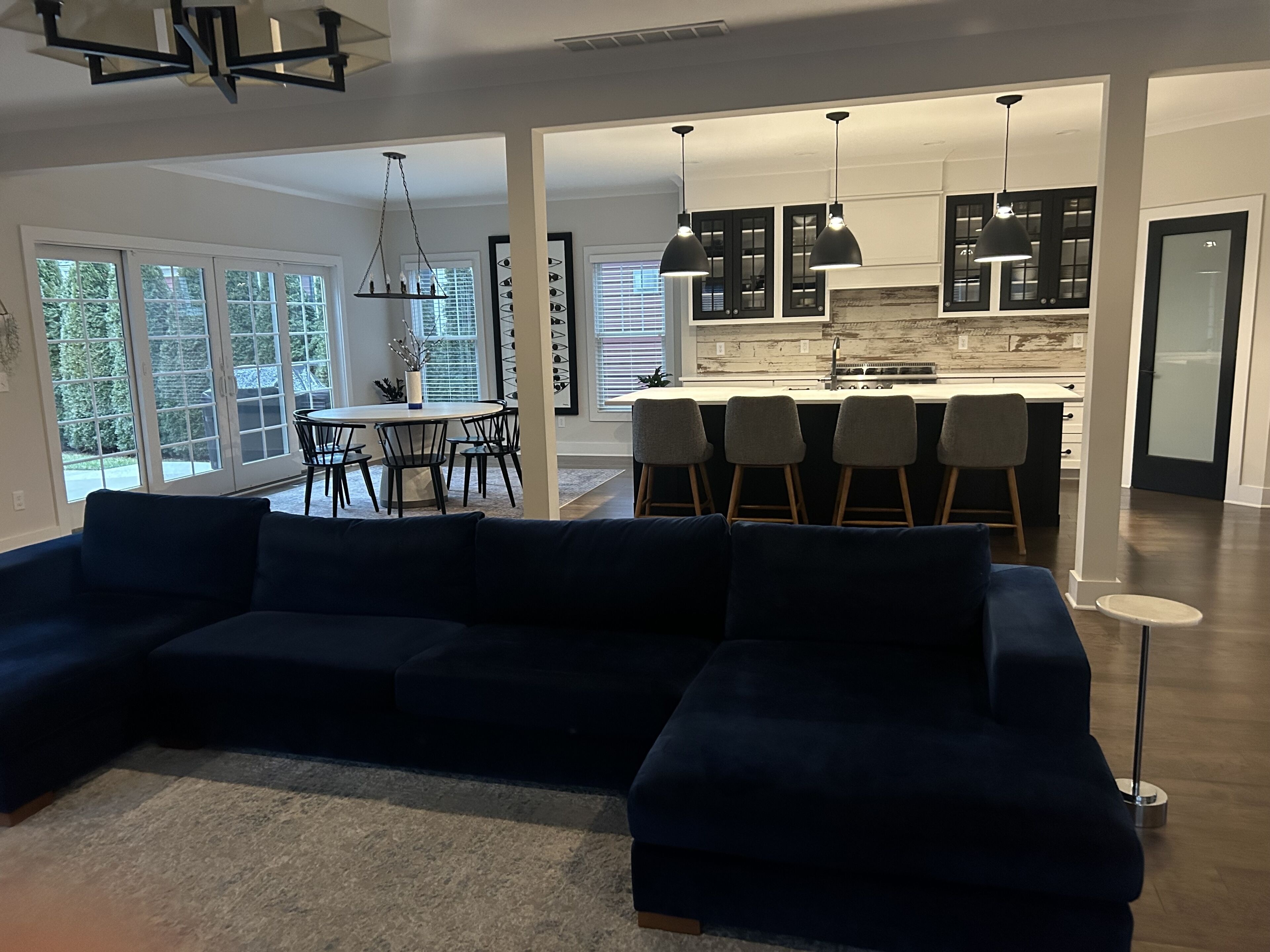 A modern living room with a dark blue sectional sofa, a dining area with a round table and chairs, and a kitchen with dark cabinets and pendant lighting.