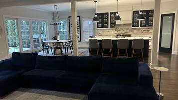 A modern living room with a dark blue sectional sofa, a dining area with a round table and chairs, and a kitchen with dark cabinets and pendant lighting.