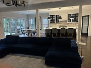 A modern living room with a dark blue sectional sofa, a dining area with a round table and chairs, and a kitchen with dark cabinets and pendant lighting. - Notre Dame Ave. Dream Home Built in 2018 -1.5 Miles from Campus (South Bend)