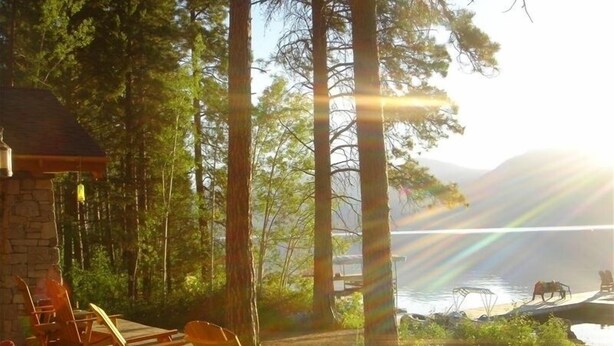 A lakeside patio with wooden chairs and a stone table, surrounded by tall trees and a view of the lake. - The Most Beautiful House on Flathead Lake - NW Travel Magazine 'Best Waterfront' (Polson)