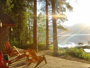 A lakeside patio with wooden chairs and a stone table, surrounded by tall trees and a view of the lake. - The Most Beautiful House on Flathead Lake - NW Travel Magazine 'Best Waterfront' (Polson)