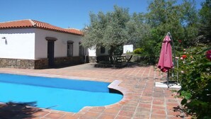 Una piscina con bordo piastrellato, un edificio bianco con tetto in tegole rosse e un ombrellone rosa.