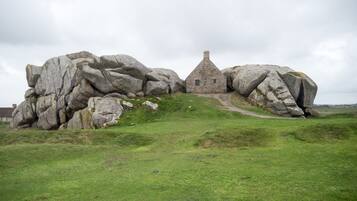 A stone house with a thatched roof nestled between large boulders on a grassy hill.