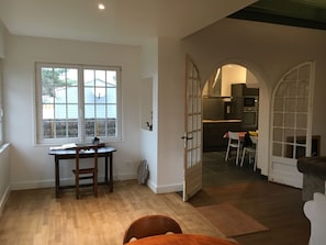 A spacious room with a wooden floor, a dining table with chairs, a window with blinds, and an open door leading to a kitchen area.