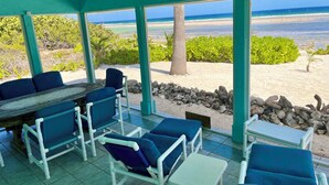 Ein überdachter Außenbereich mit blauen, gepolsterten Stühlen und einem Tisch, mit Blick auf einen Strand mit klarem, blauem Wasser und sandigem Ufer.