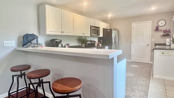 A kitchen with white cabinets, a microwave, a refrigerator, a coffee maker, and a sign that says 'HELLO'. There are three stools and a small island with a sink.
