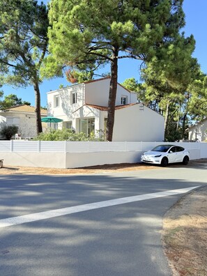 Une maison blanche à deux étages, avec un toit recouvert de tuiles rouges, une clôture blanche et une voiture blanche garée devant.