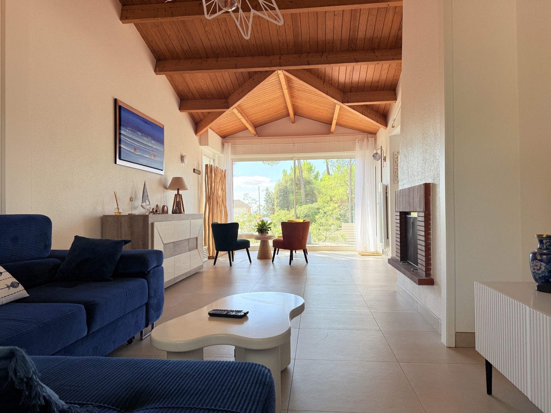 A modern living room with a sofa, coffee table, and chairs, a fireplace, and a large window with a view of greenery.