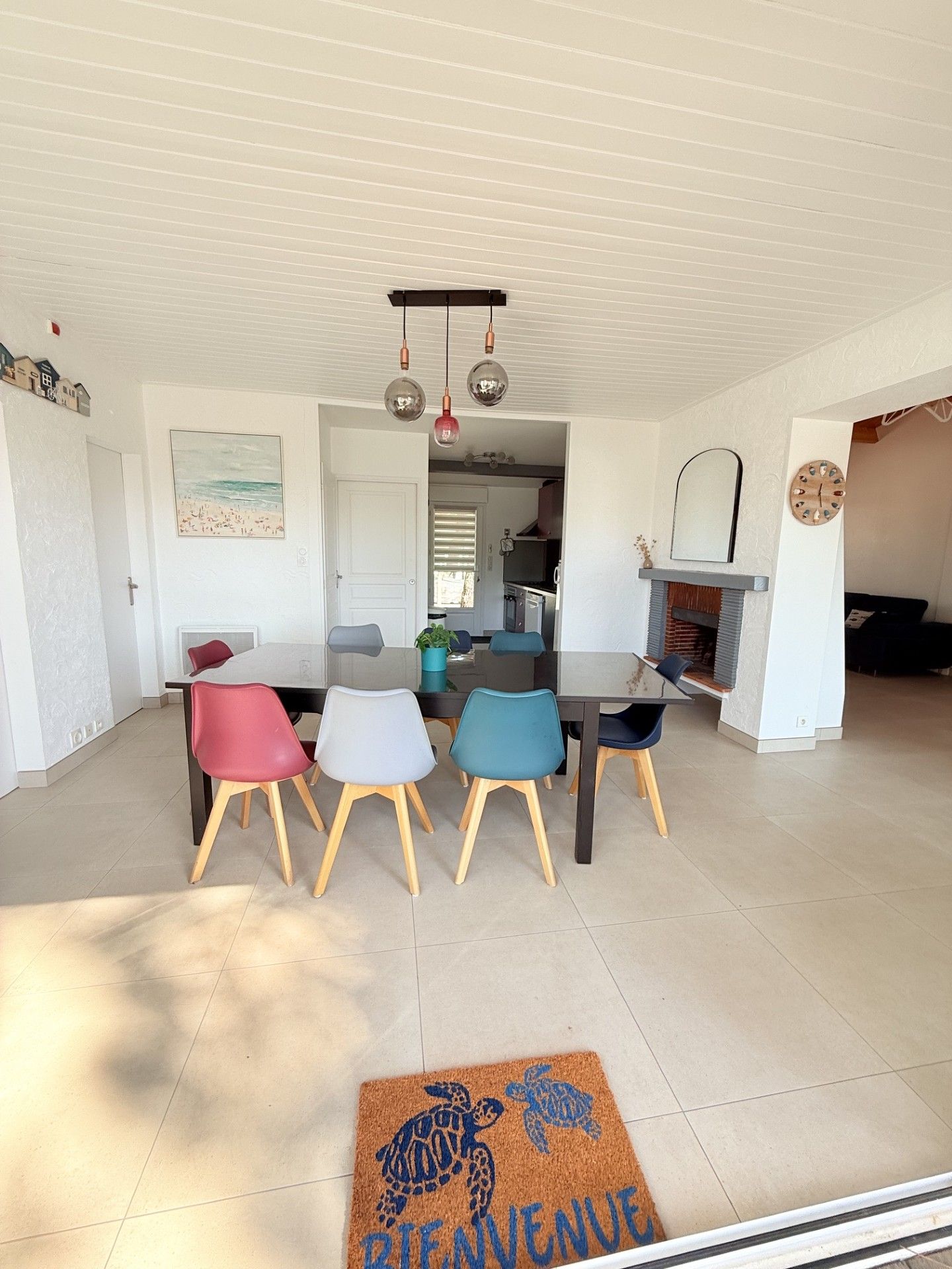 A dining area with a table and chairs, a kitchen in the background, and a welcome mat with a turtle design.