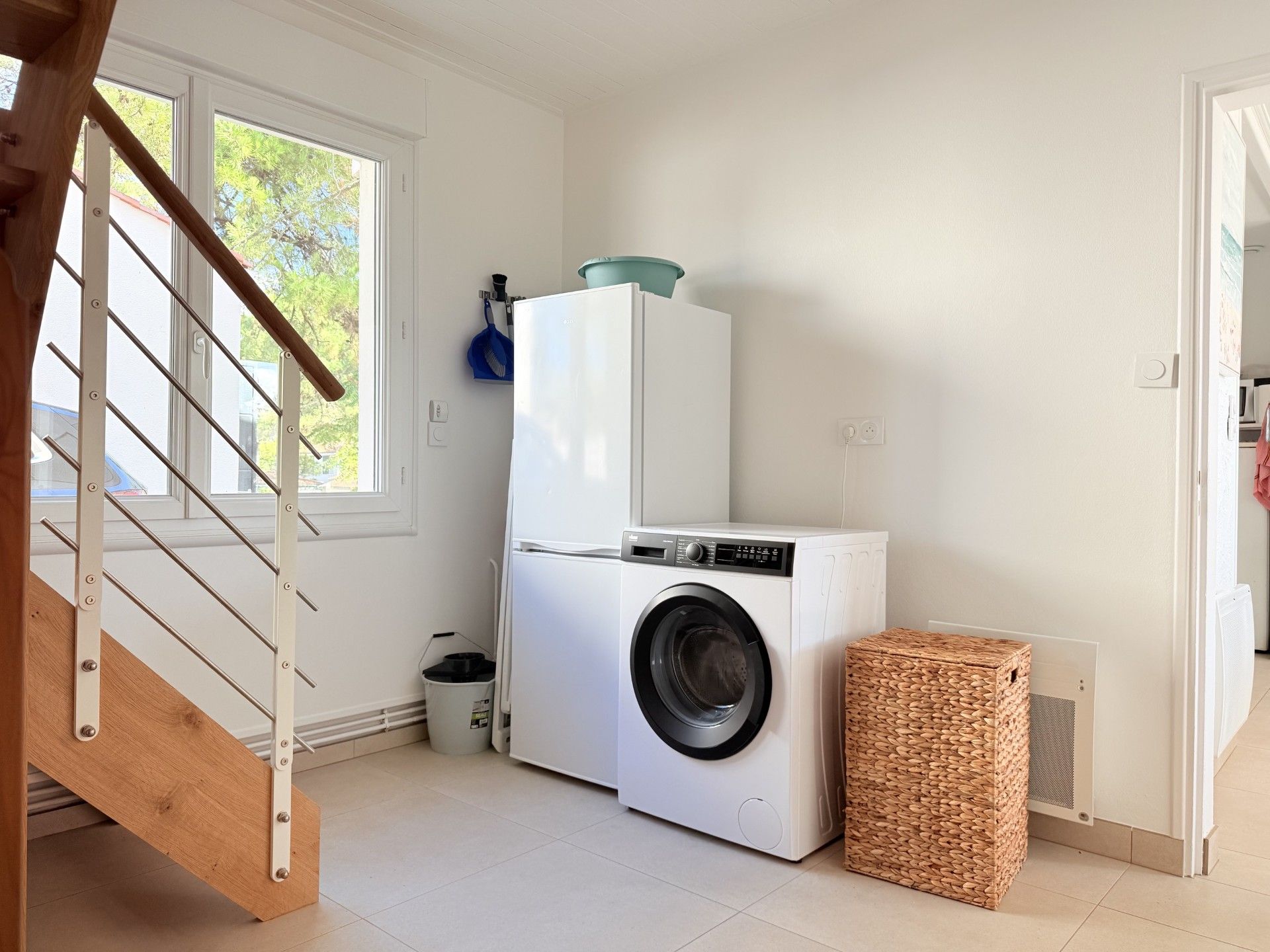 A laundry room with a washing machine and dryer, a wooden staircase, and a window with a view of trees.
