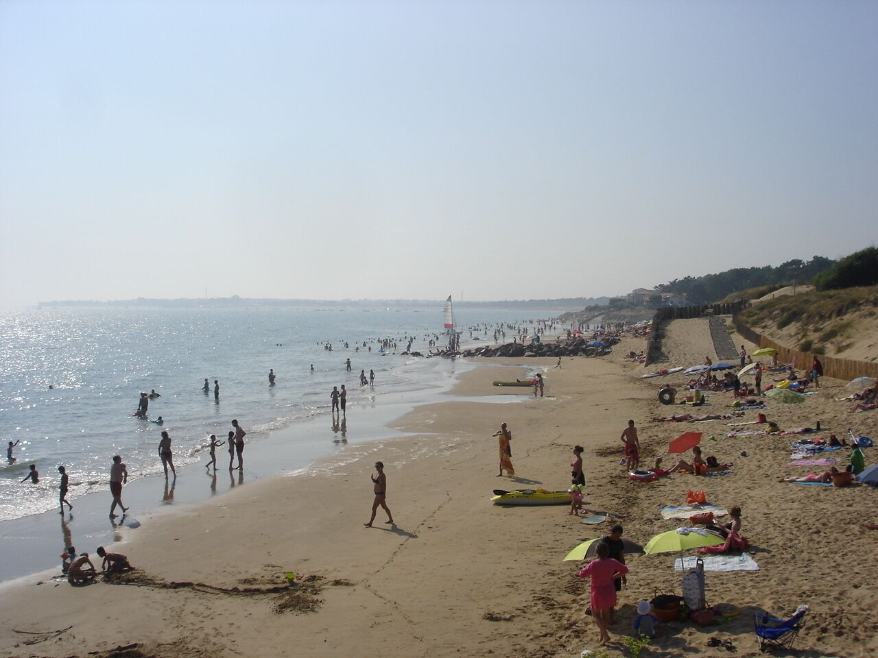 Una playa concurrida con gente caminando y nadando, y un velero a lo lejos.