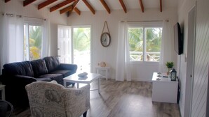 A spacious living area with a dark leather sofa, a white coffee table, and a patterned armchair. A small white side table is near the sofa. The room has wooden beams on the ceiling and large windows with curtains.