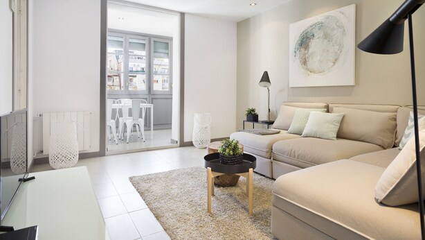 A modern living room with a sofa, a coffee table, and a floor lamp. There is a large window and a door leading to an outdoor area.
