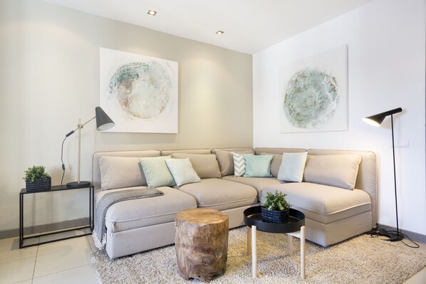 A modern living room with a beige sectional sofa, a round wooden coffee table, a black side table with a lamp, and abstract wall art.