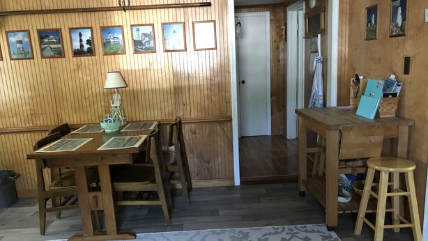 Een kamer met houten lambrisering, een eettafel, stoelen, een klein krukje, een lamp en een vloerkleed.