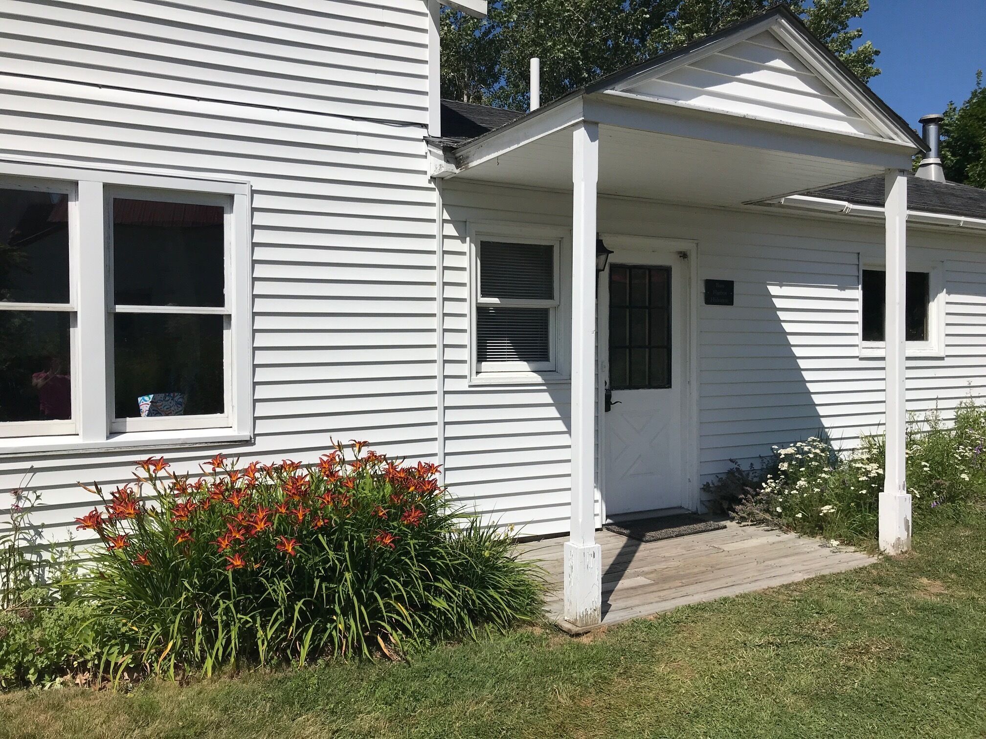 A white single-story house with a front porch, a small garden with red flowers, and a clear sky.