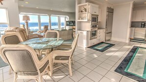 Un'ampia sala da pranzo con tavolo in vetro circondato da sedie in vimini, una cucina con mobili bianchi e vista sull'oceano attraverso ampie finestre.