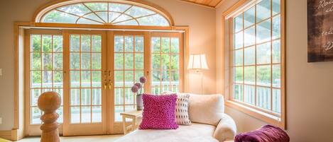 A cozy bedroom with a large window, a wooden ceiling, and a view of the outdoors.
