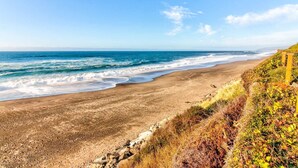 Una spiaggia con onde, cielo sereno e un promontorio roccioso con vegetazione.