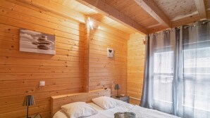 A wooden cabin bedroom with a bed, pillows, a nightstand, a lamp, a picture of stones, and a window with curtains. - CHALET RIVIERA "JACUZZI OFFERT" 3* wifi ANDORRA ax les thermes Orlu ski (Orlu)