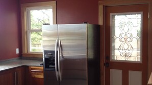 A kitchen with a stainless steel refrigerator, wooden cabinets, a window with a view, and a door with a decorative glass panel. - Minnesota Point Beach House 1-Spectacular Lake Superior View (Duluth)