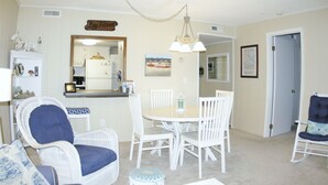 Un coin repas avec une table ronde et des chaises blanches, un fauteuil en osier avec un coussin bleu et un lustre.