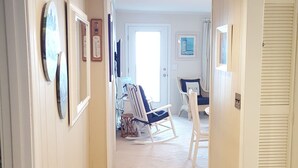 Un couloir doté d’un plancher en bois, d’un fauteuil à bascule, d’une chaise avec un coussin portant l’inscription « RELAX » et d’une porte avec un volet.