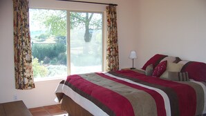 A bedroom with a large bed, floral curtains, a window with a view of greenery, and a tiled floor. - Lupine Ranch - Wine Country Guest House (Templeton)