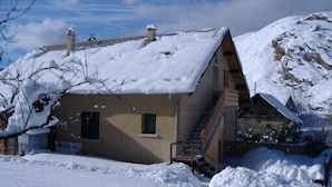 A snow-covered building with a sloped roof, a small window, and a staircase leading to a wooden deck. - Apartment in a quiet village (Briançon)
