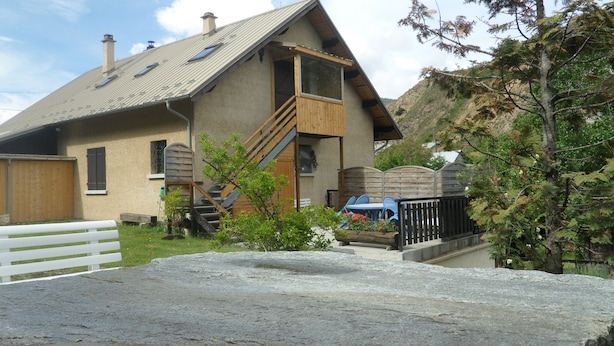 A rustic house with a wooden staircase leading to a balcony, a fenced yard, and a vehicle parked outside. - Apartment in a quiet village (Briançon)