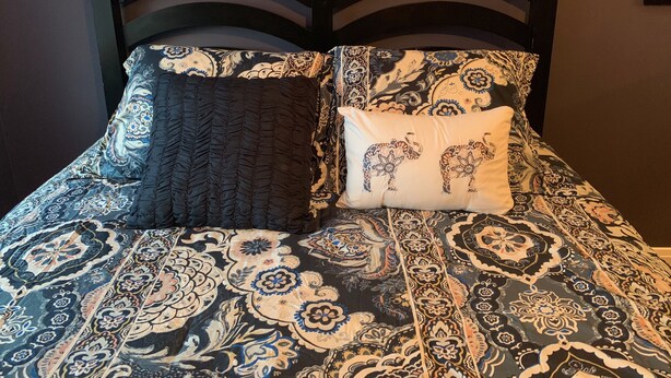 Un lit avec une couvre-lit à motifs et deux oreillers, l’un recouvert d’un tissu foncé et l’autre orné d’un motif d’éléphant.